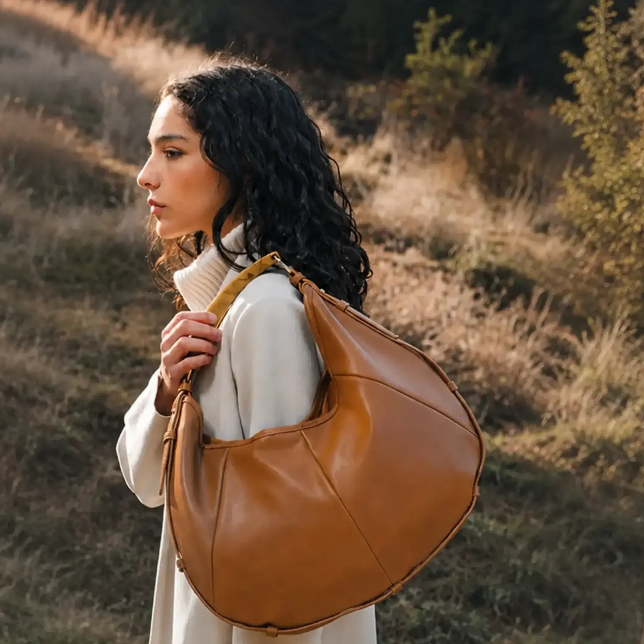 A woman in a long cream coat stands on a grassy hillside at golden hour, carrying a large tan leather hobo shoulder bag.