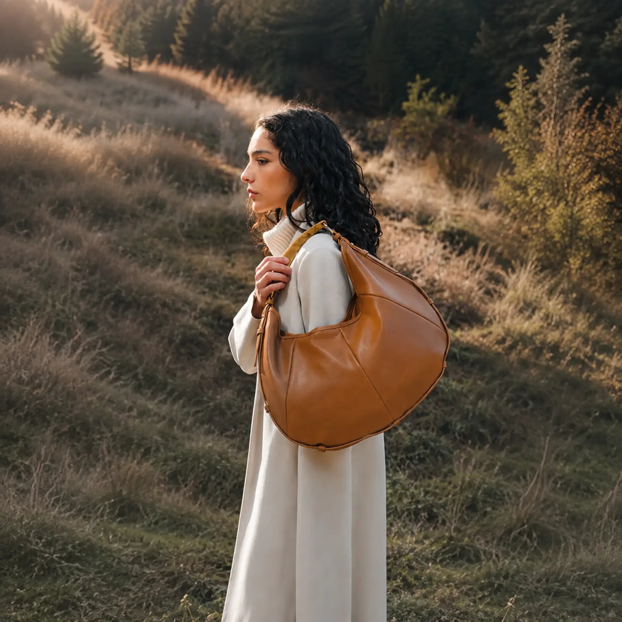 A woman in a long cream coat stands on a grassy hillside at golden hour, carrying a large tan leather hobo shoulder bag.