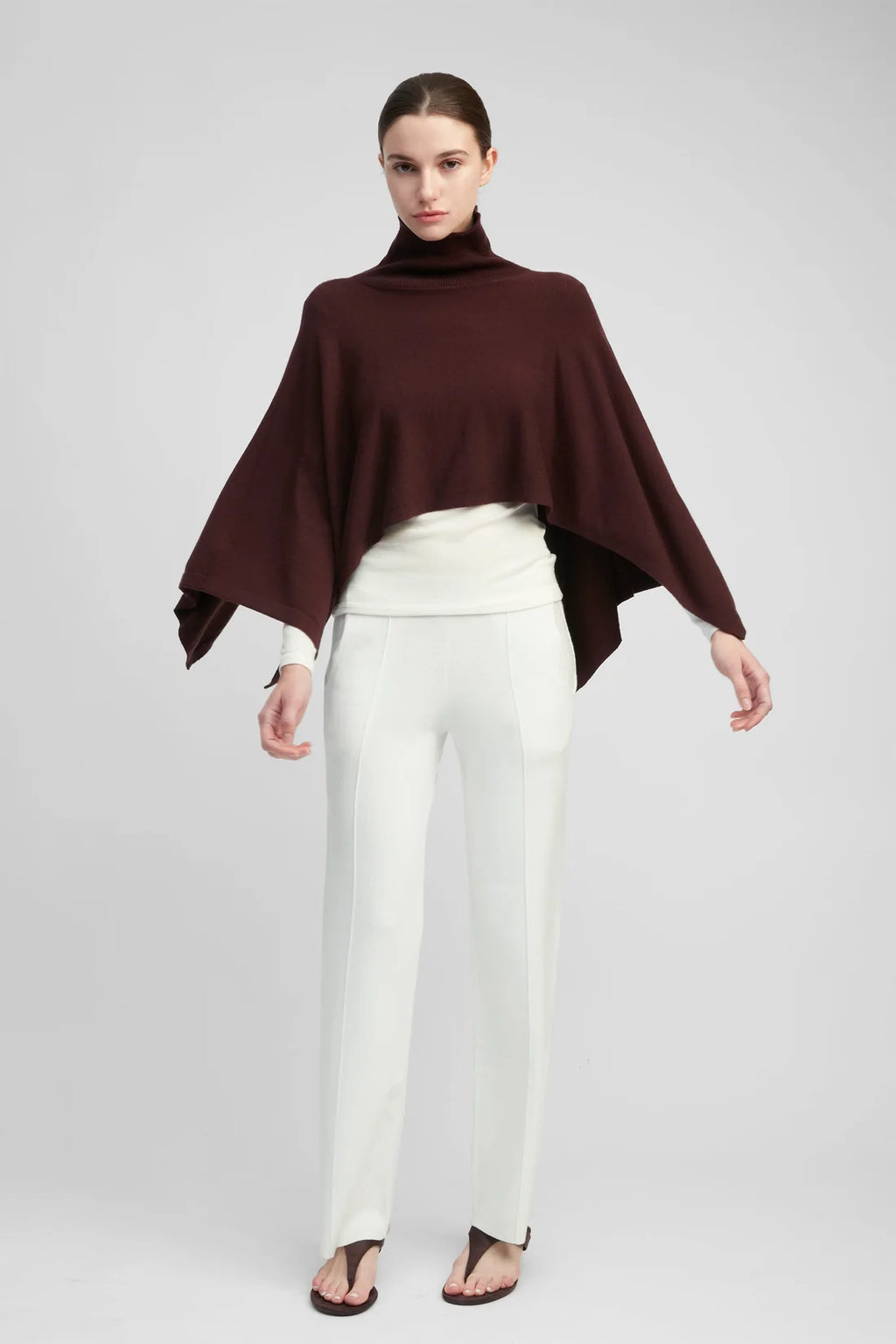 Woman wearing a burgundy poncho and white pants on a gray background