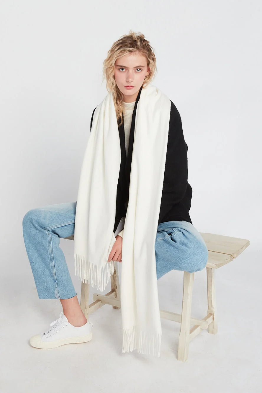 Model wearing an ivory cashmere-feel oversized women's scarf with fringe detailing, styled long over a black jacket.