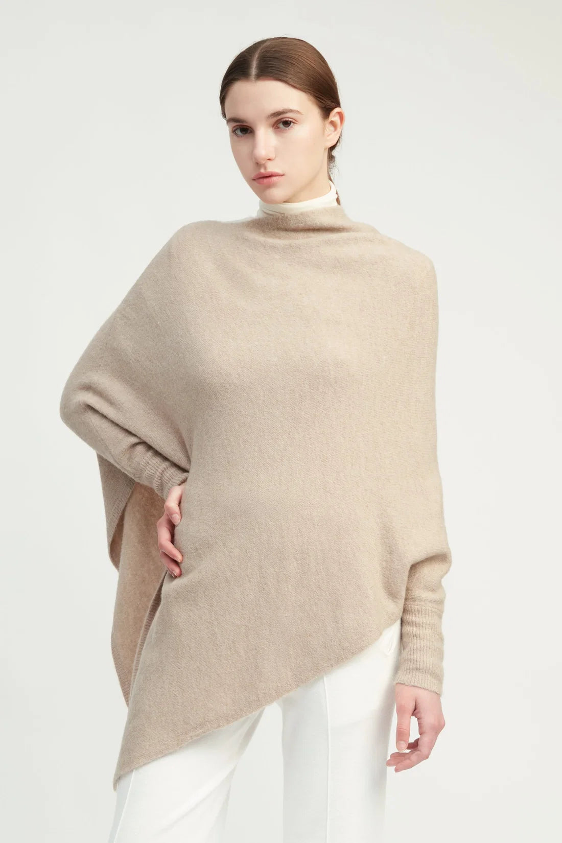 A woman modeling a light beige knit poncho sweater over a white top and white pants, photographed against a clean light gray studio background.
