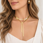 A close-up of a woman wearing a minimalist gold double-loop lariat necklace with a long pendant drop, styled with a white top and a gold hoop earring.