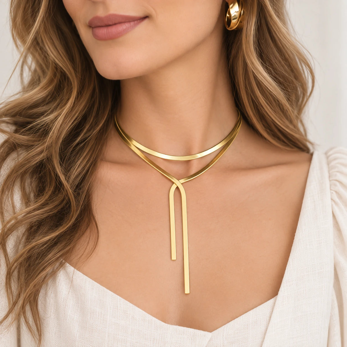 A close-up of a woman wearing a minimalist gold double-loop lariat necklace with a long pendant drop, styled with a white top and a gold hoop earring.