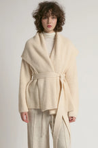 A model wearing a cream-colored ribbed knit belted wrap jacket over a white turtleneck and matching cream trousers, set against a plain light gray studio background.