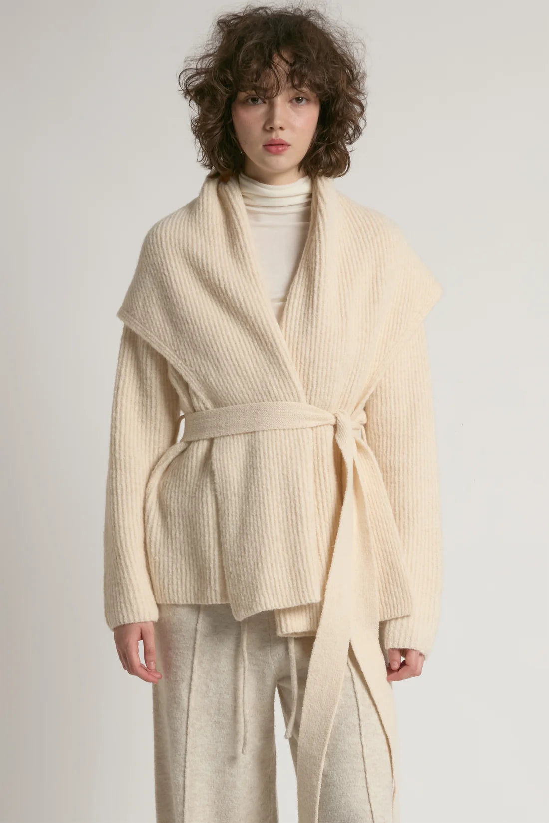 A model wearing a cream-colored ribbed knit belted wrap jacket over a white turtleneck and matching cream trousers, set against a plain light gray studio background.