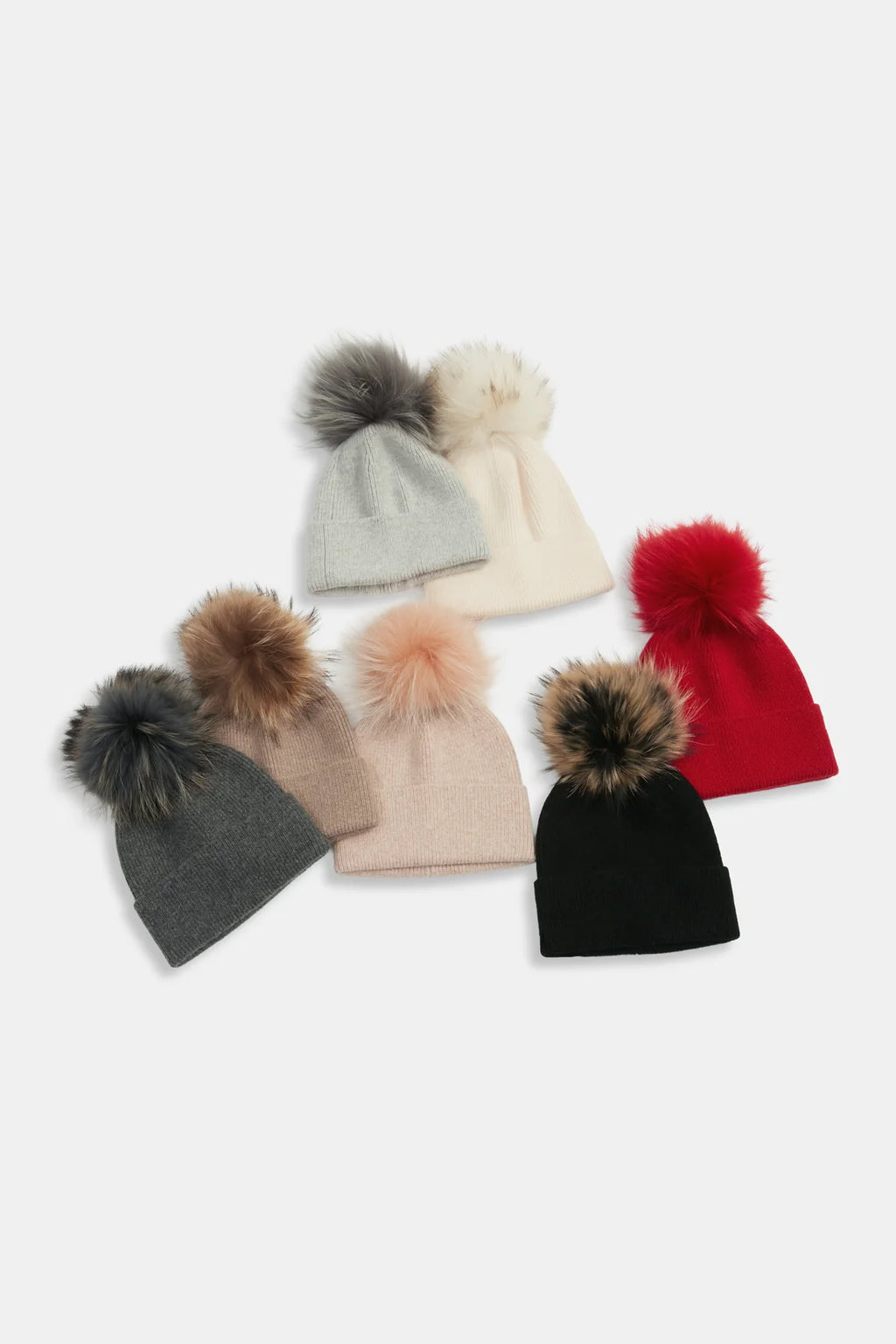 Six knit beanies with large faux-fur pom-poms arranged on a light gray background. The beanies are in gray, cream, beige, red, black, and blush pink, with the pom-poms in matching or contrasting tones.