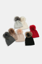 Six knit beanies with large faux-fur pom-poms arranged on a light gray background. The beanies are in gray, cream, beige, red, black, and blush pink, with the pom-poms in matching or contrasting tones.