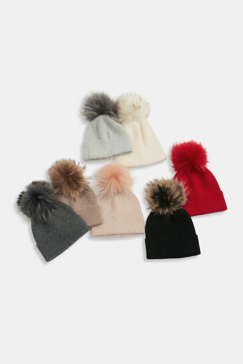 Six knit beanies with large faux-fur pom-poms arranged on a light gray background. The beanies are in gray, cream, beige, red, black, and blush pink, with the pom-poms in matching or contrasting tones.